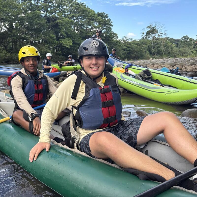 Two young men are sitting in an inflatable raft on a river, wearing life jackets and helmets. They appear to be preparing for or just finishing a rafting trip. Other rafts and people are visible in the background, along with lush green trees lining the riverbank. The sky is partly cloudy, suggesting a bright day for outdoor activities.